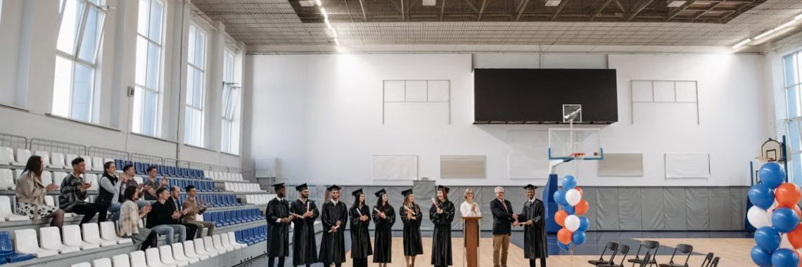 A graduation ceremony held in an indoor gymnasium with graduates and an audience.